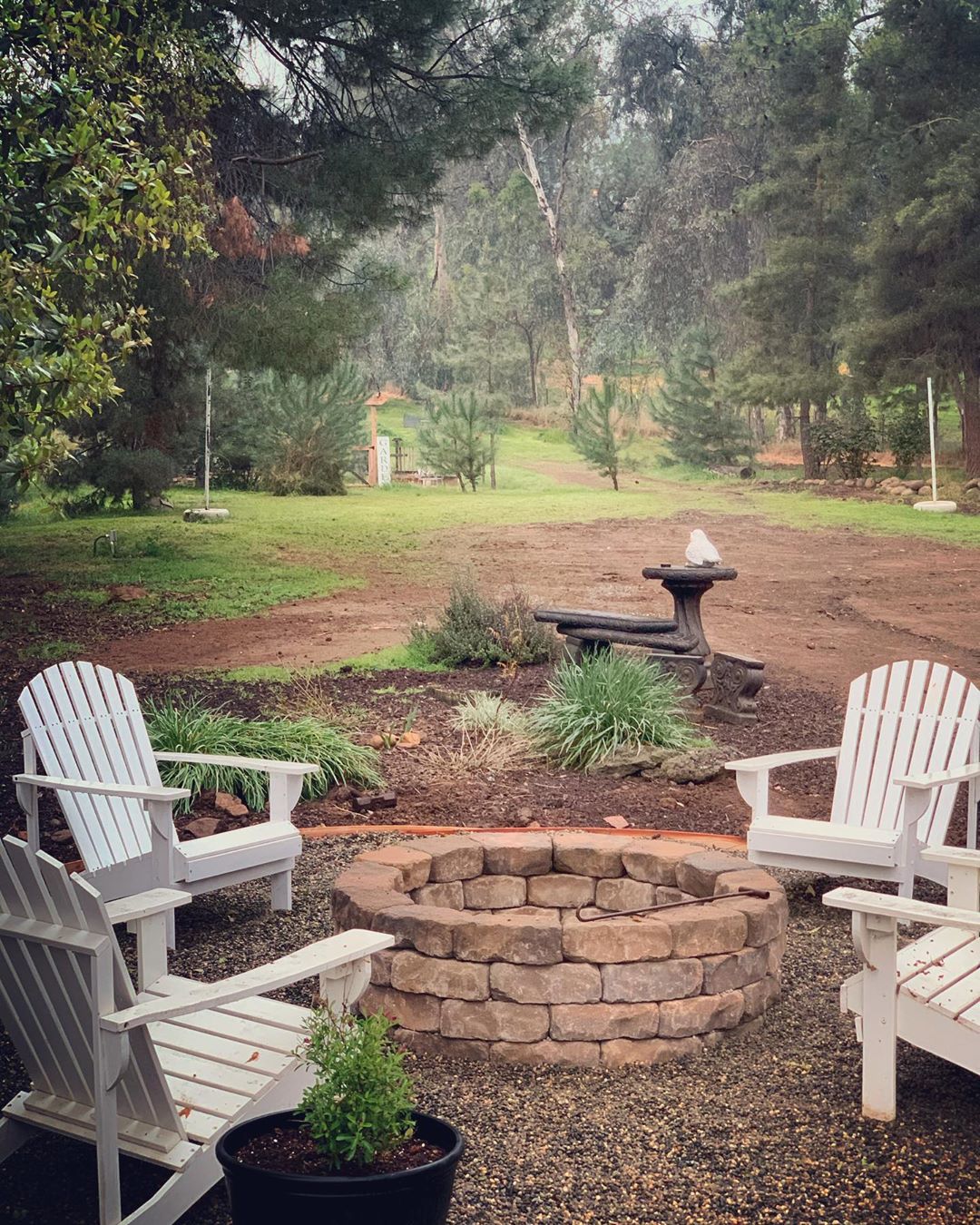 backyard fire pit built with large stones and white adirondack chairs around photo by Instagram user @claudio_paisagismo