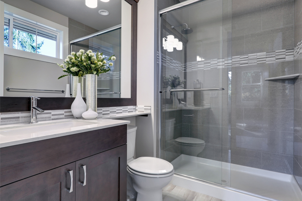 A gray toned bathroom with a glass door shower and a big mirror behind the sink vanity and toilet