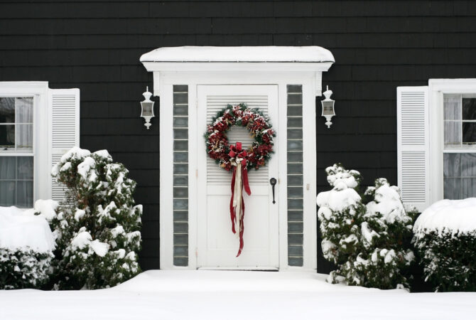 A white door with a traditional pine wreath and red and gold ornaments is hanged on a white door of a house surrounded by snow
