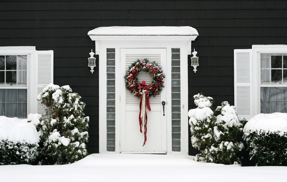A white door with a traditional pine wreath and red and gold ornaments is hanged on a white door of a house surrounded by snow