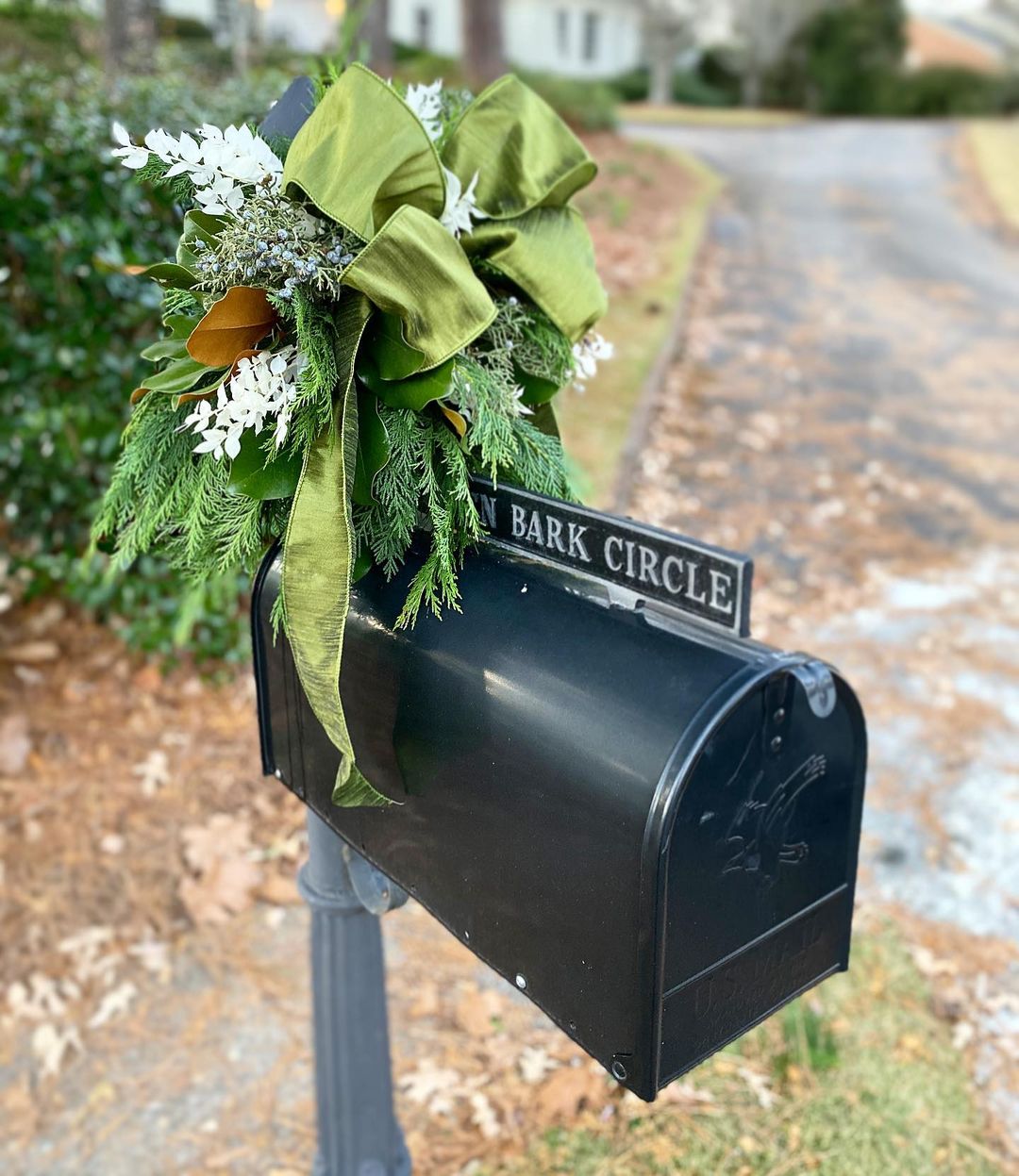 A black mailbox is decorated with a green bow, evergreen sprigs, juniper sprigs and berries, and white flowers. Photo by Instagram user @wildthingsbirmingham.
