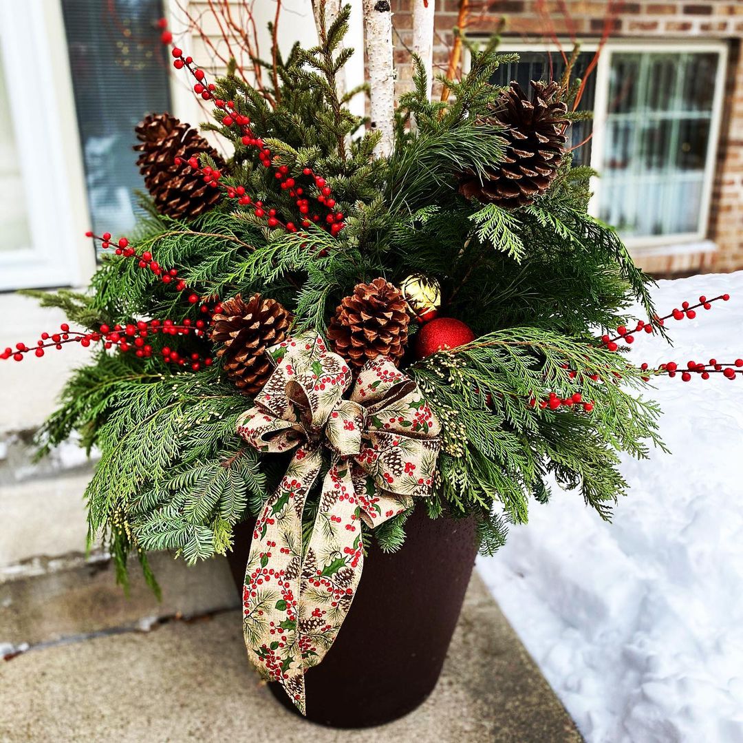 Front porch planter stuffed with pine sprigs, evergreen sprigs, cranberry shoots, pinecones, a large bow, and ornaments. Photo by Instagram user @junealltrades. 