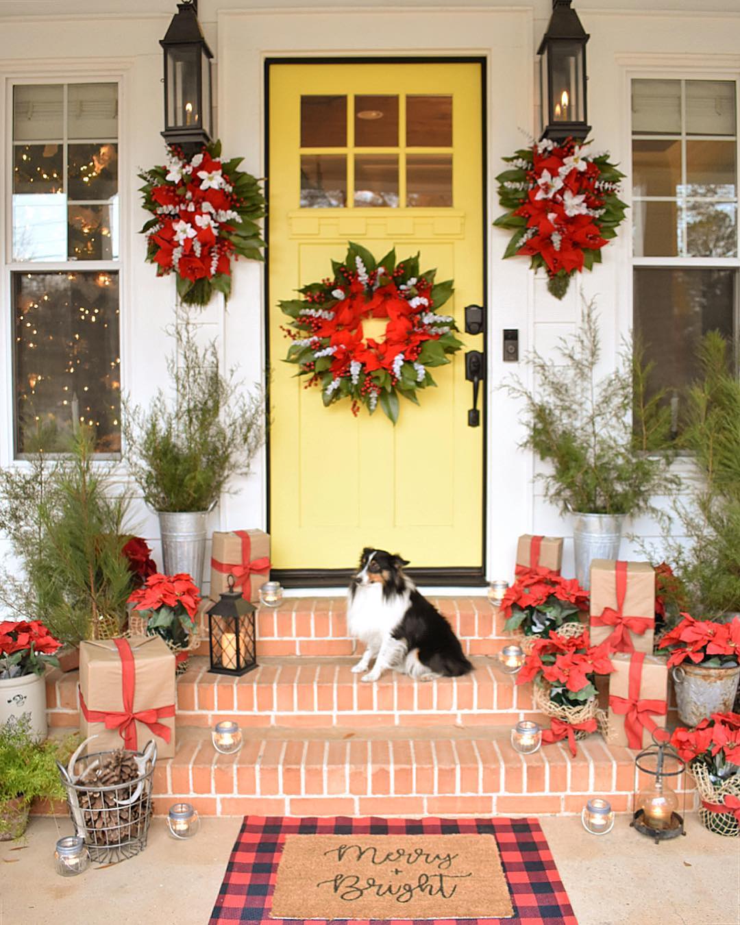 Yellow front door features a poinsetta and pine wreath, and two additional poinsettia wreathes hang from porch lanterns on either side of the door. A dog is sitting on the steps between various other planters with pine- and holiday-related decorations. Photo by Instagram user @simplysoutherncottage.