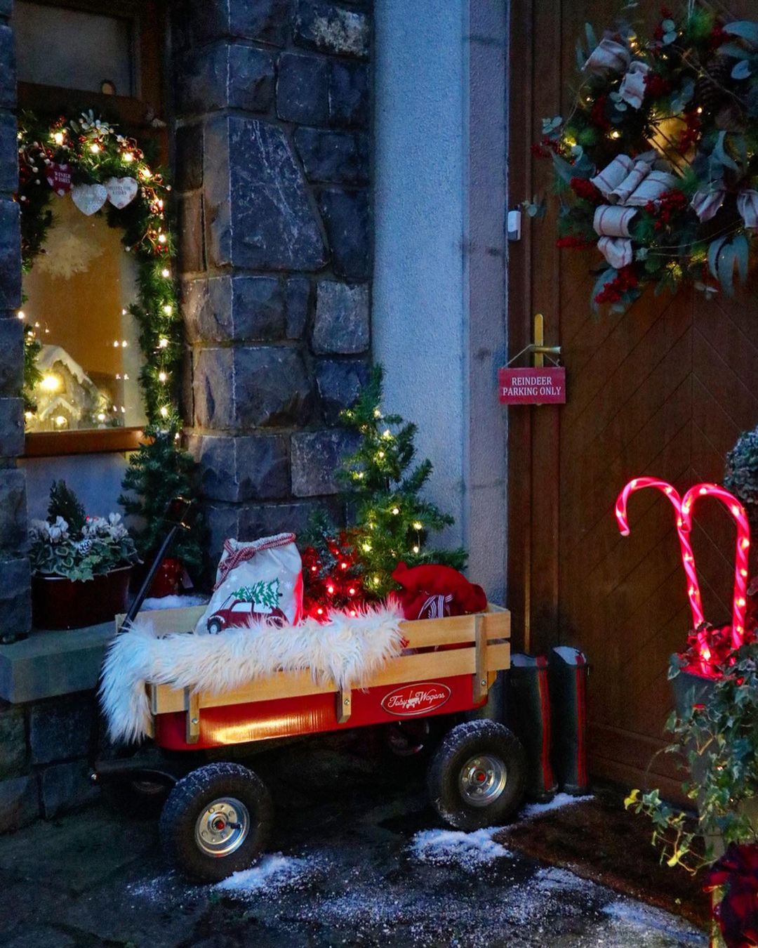 A small red and wooden wagon sits on the front porch holding a mini, lit Christmas tree, cotton material, and other various holiday decor. Photo by Instagram user @my_irish_home. 