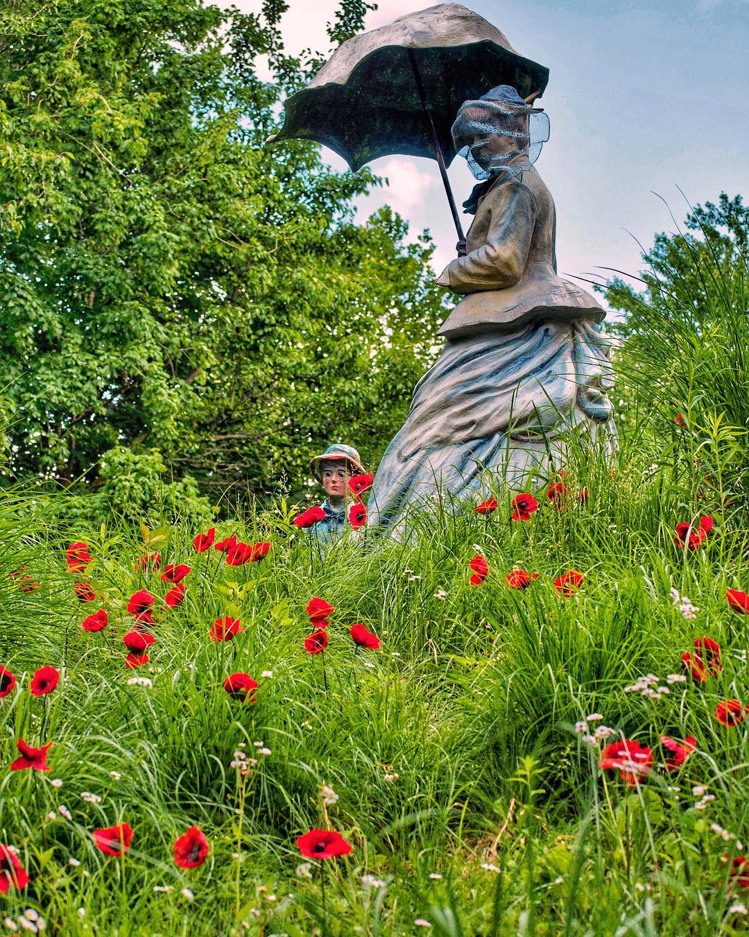 Statue "On Poppied Hill" by Seward Johnson in Grounds for Sculpture, NJ. Poppies are red and in full bloom amongst long grass and tiny purple flowers. Statue is of a woman in the Victorian era, holding a sun parasol. Photo by Instagram user @thedailyantiquarian