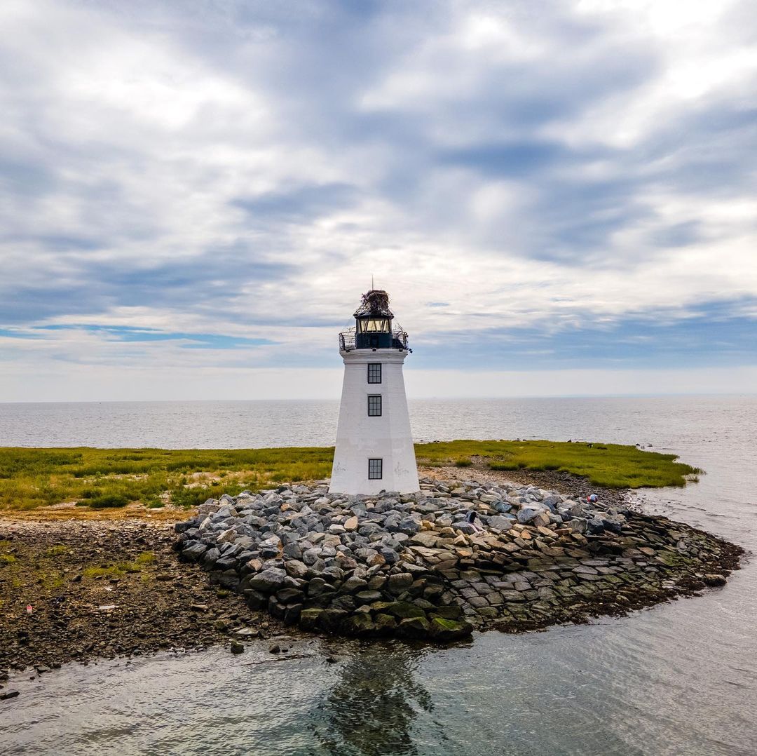 Fayerweather Lighthouse in Bridgeport, CT: mid-size lighthouse on short rocky outcropping on bright cloudy day in Bridgeport. Photo by @marylanddroneguy.