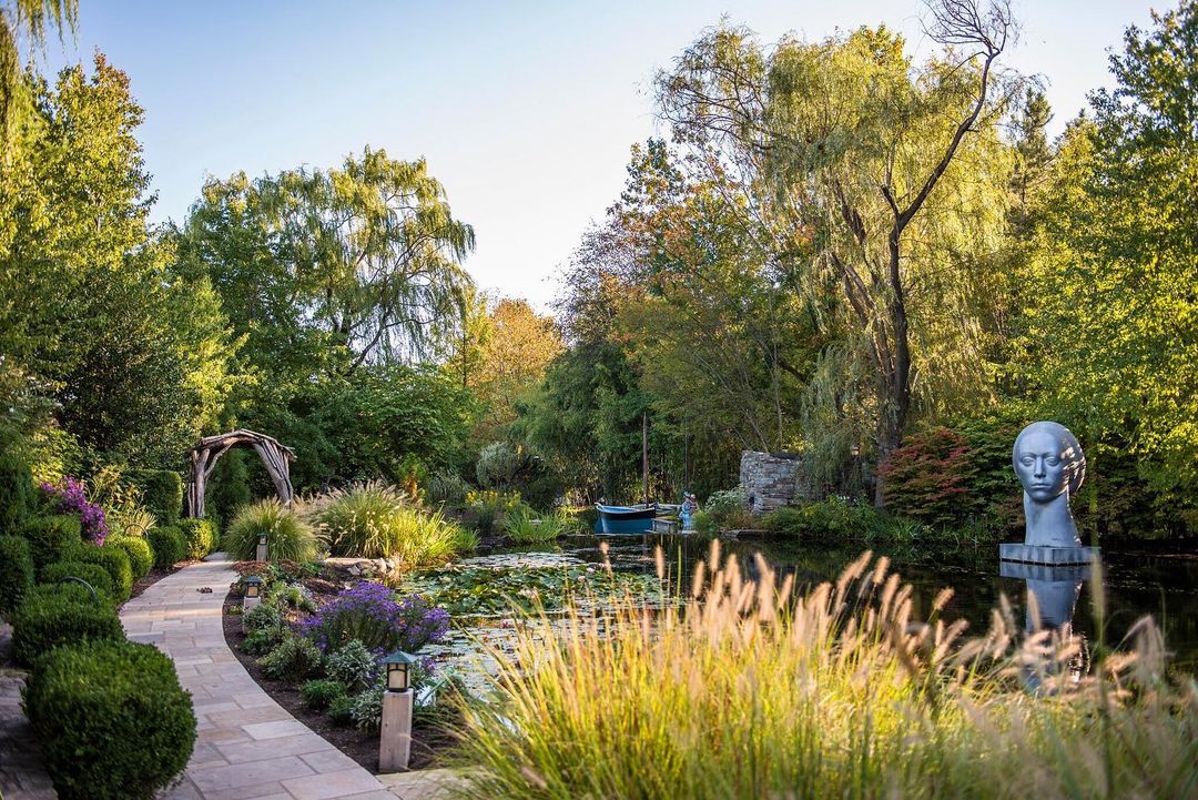 Shot of Grounds for Sculpture featuring several species of trees, bushes, and flowers, as well as water plants. A striking statue of a head rises above the water. Photo by Instagram user @groundsforsculpture