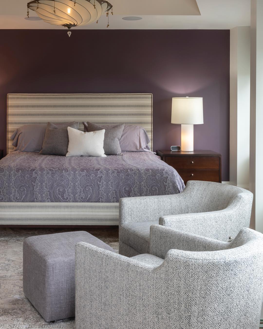 Plum accent wall in a room with a lavender bed and light gray chairs.