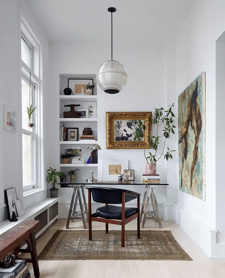 Interior of white-walled home office with Mid-Century Modern black leather chair and glass desk surrounded by antique paintings and greenery