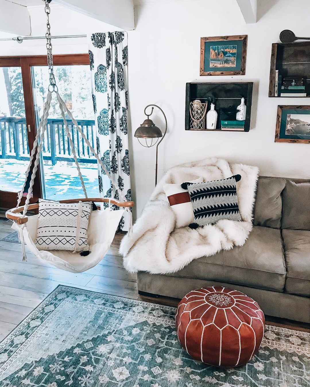 Living room with cool touches, including a dark teal rug, teal and navy-patterned curtains, and soft grey sofa, beside a hanging chair made of wood, white canvas material. Photo by Instagram user @shoppingwith.hannah.