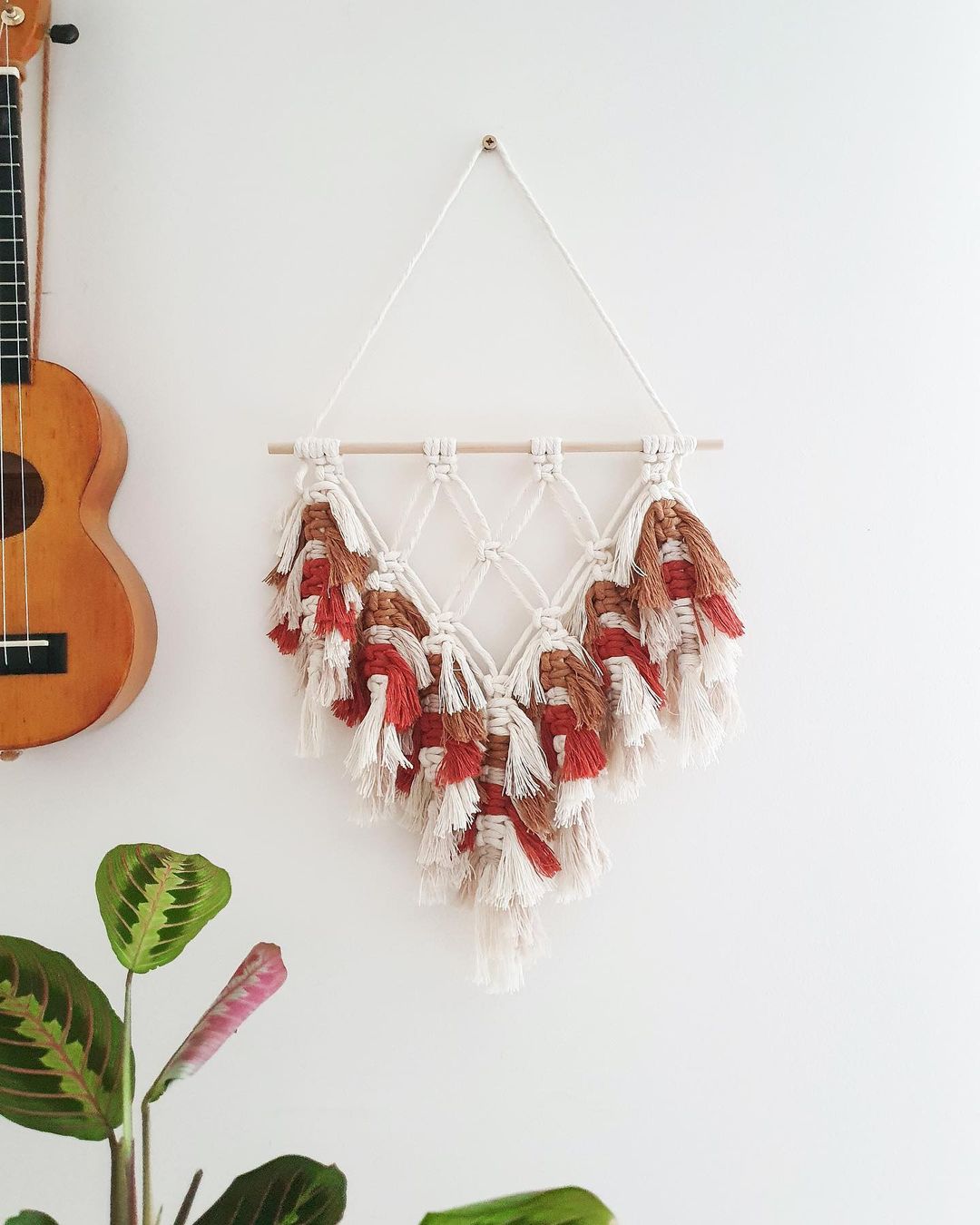 Wall with a ukelele, prayer plant, and tan,rust, and white macrame art piece. Photo by Instagram user @cozyknotsdesign.