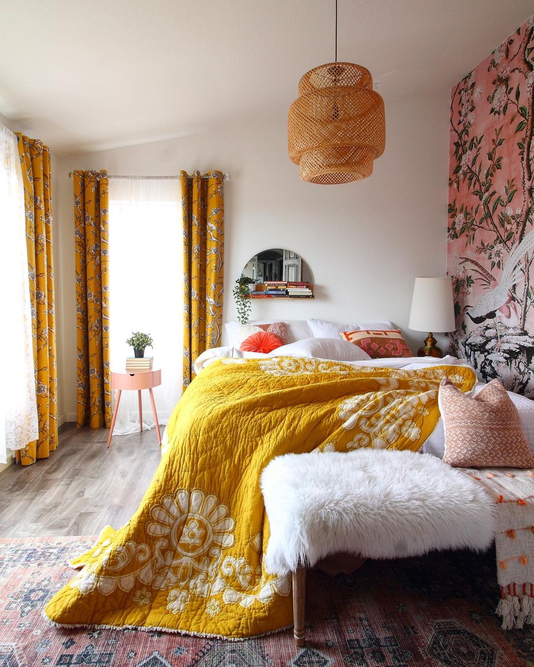 Bedroom with large windows, a lamp to the left of the bed and a hanging light with a wicker shade. The room rug has a faded, antique effect on an aztec design, and an accent wall with a scene of two birds amidst a pink sky and gentle, budding branches of a flowering tree. Photo by Instagram user @raveinteriordesign.