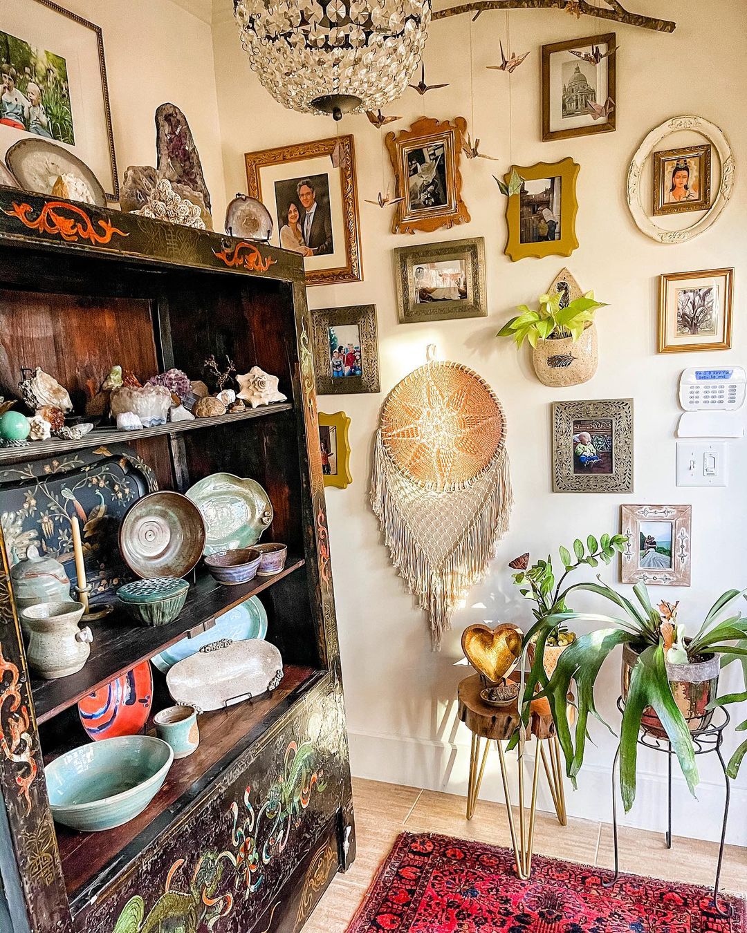 Decorated, antique bookshelf presenting vintage platters, pots, shells, and geods, beside plants and a wall with photos framed in old, unique frames. Photo by Instagram user @the.rustic.bohemian.