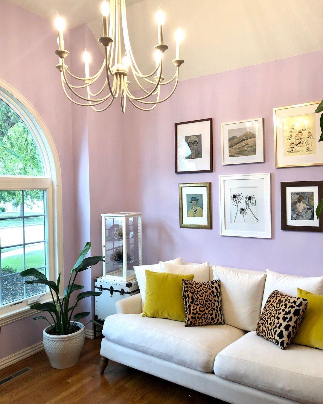 Sitting area with a lilac purple wall next to a bay window and potted plant with artwork on the wall. Photo by instagram user @sugarkanedesigns