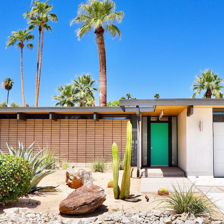 Exterior of a modern home in Palm Springs with a desert landscaped front yard and palm trees in the background. Photo by instagram user @modtraveler