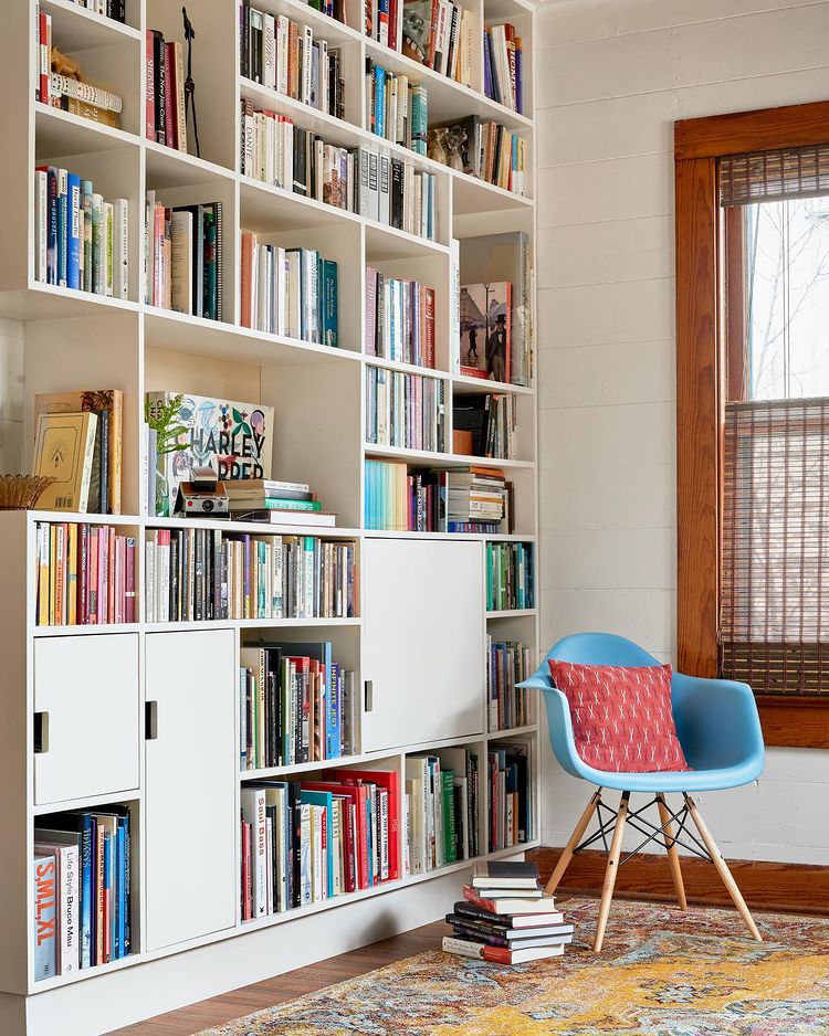 Modern home library with built-in white shelving holding many books with a blue statement chair next to it. Photo by instagram user @leonidfurmanskyphotog