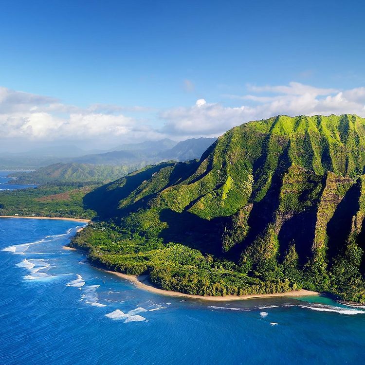 Scenic picture of Kauai, Hawaii with a sandy beach and a jungle and blue skies with clouds forming in the background. Photo by instagram user @ihsanisland