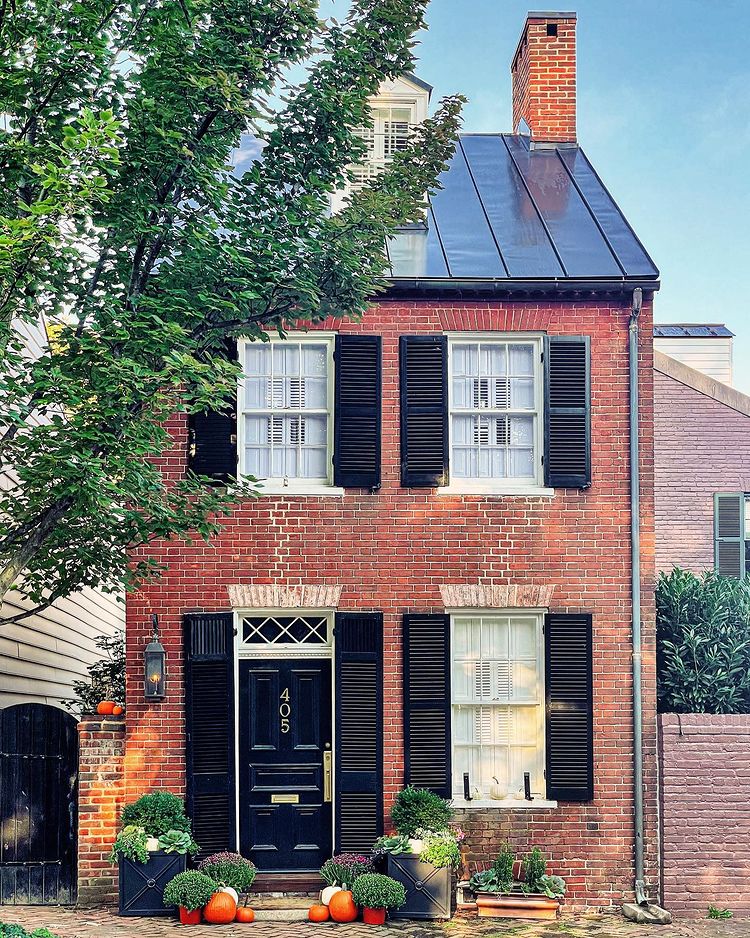 Brick colonia house with a tin roof and black shutters and a black door with pumpkins on the front porch and a gas lantern with a brick garden wall on the side. Photo by instagram user @scenefrommyrun