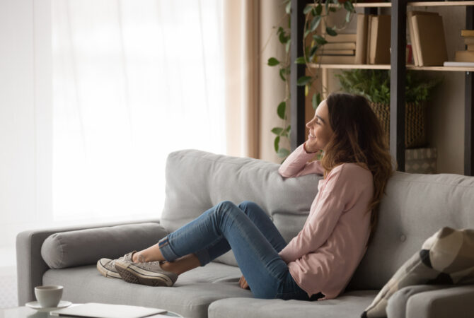 Smiling girl sitting on a comfortable couch in an apartment and looking out the window while smiling