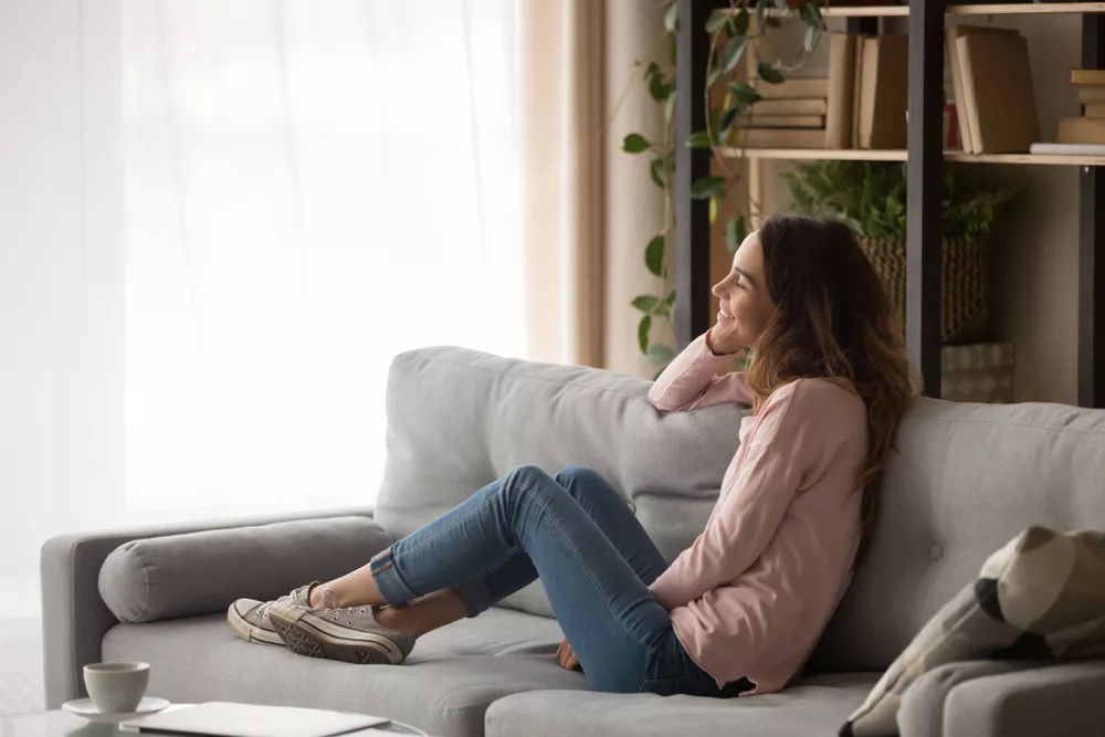 Smiling girl sitting on a comfortable couch in an apartment and looking out the window while smiling