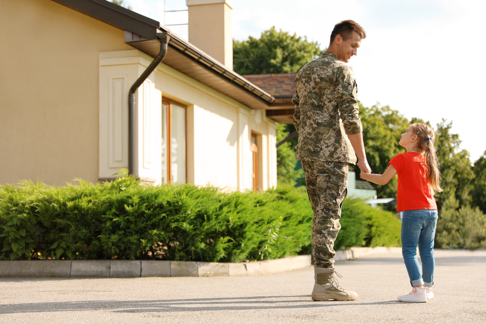 Image of a father in a military uniform holding his child's hand in front of their home.