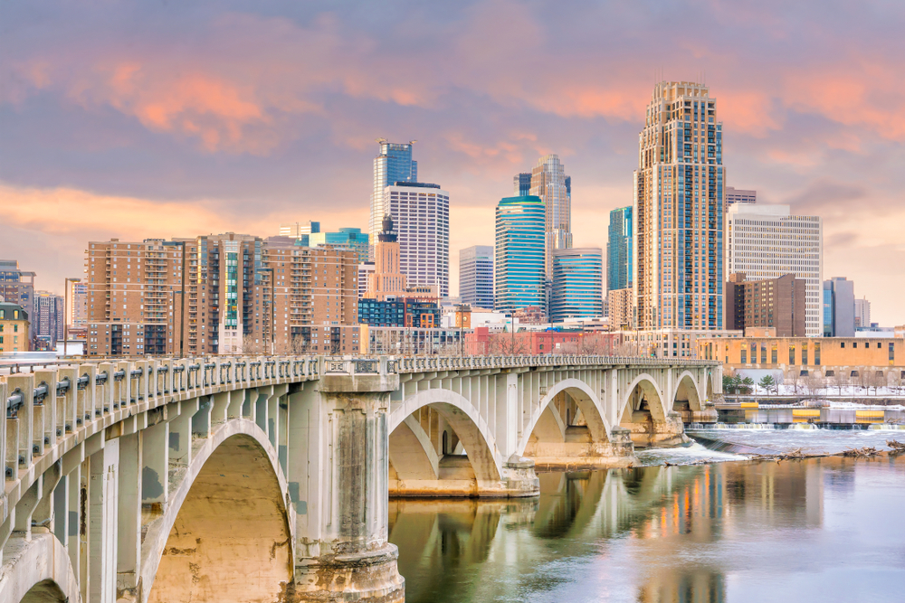 Downtown Minneapolis skyline during twilight hours with tall buildings and a beautiful bridge over a river.