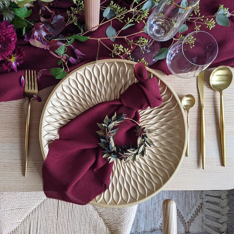 Gold and maroon christmas table with a wreath accent in the middle of the place setting. Photo by instagram user @sobremesaco