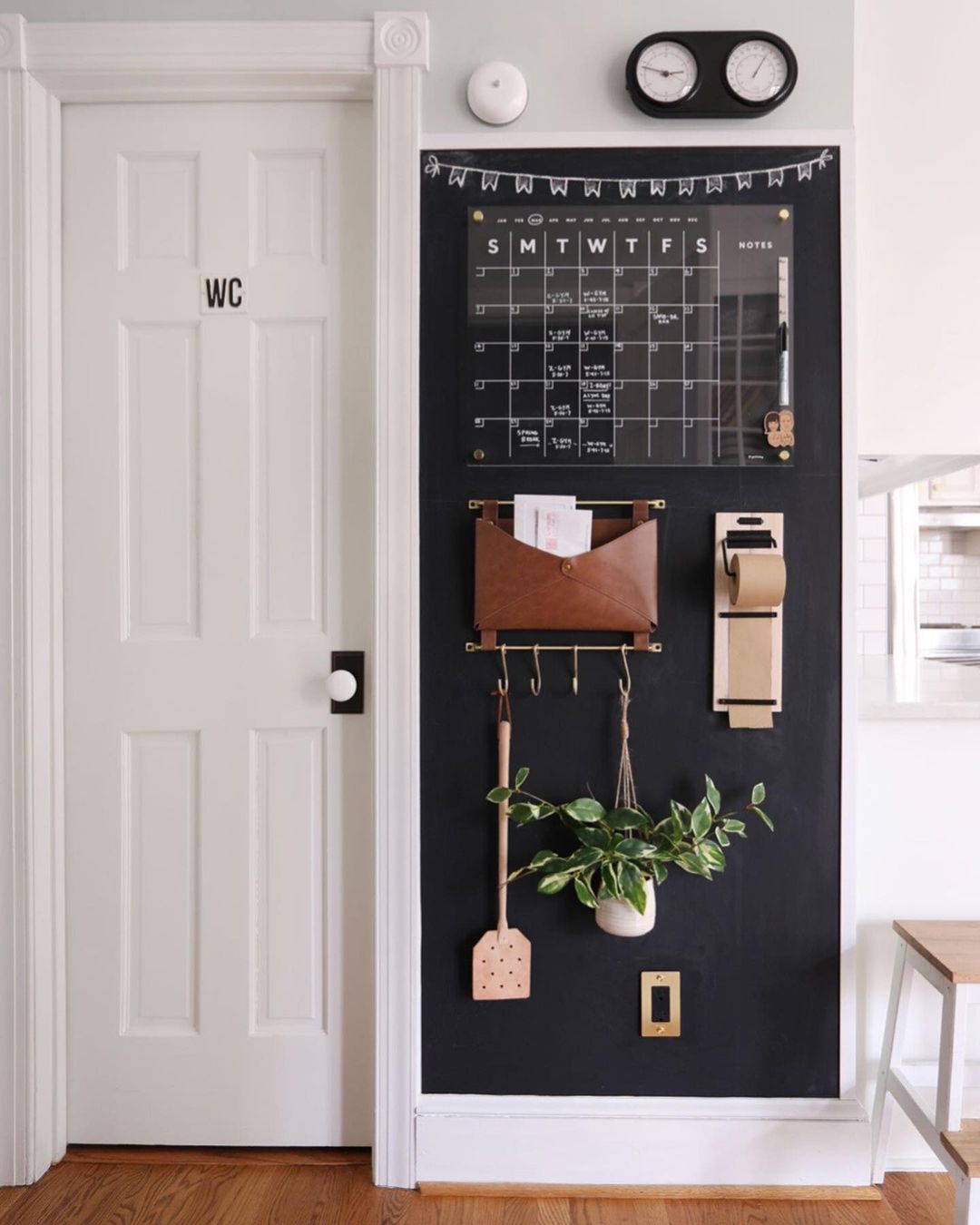 Black chalkboard accent wall next to kitchen entry with hanging acrylic calendar, cooking utensils, plant, leather pouch for envelopes and mail, and roll of brown paper for notes | Photo by Instragram user @sunnycirclestudio