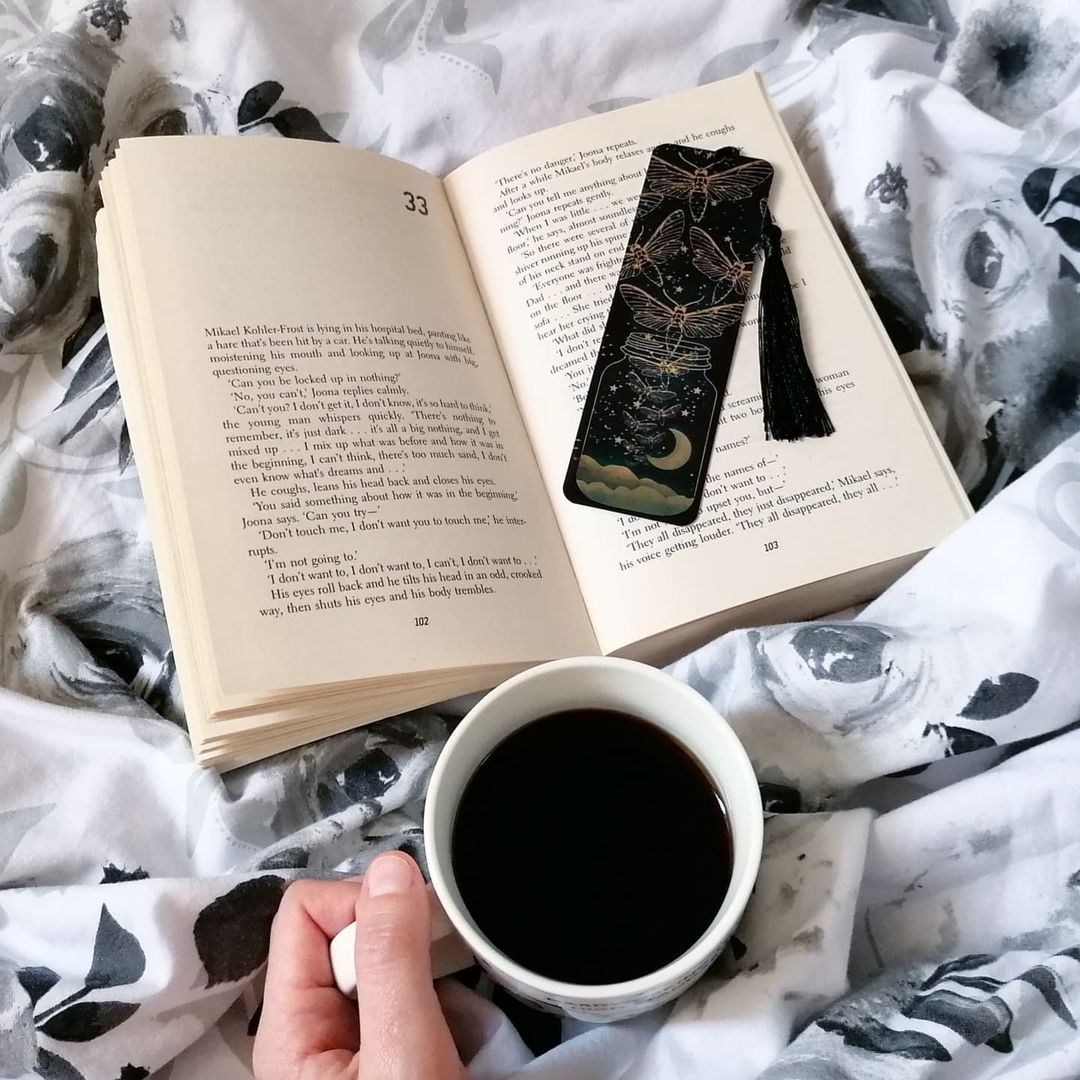Open book with firefly bookmark on black and white rose comforter next to white mug full of coffee | Photo by Instagram user @jana.reads