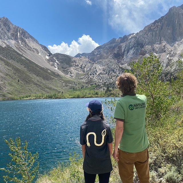 Two people with their back turned to the camera as they admire the Sierra Mountains and freshwater spring in the background | Photo by Instagram user @natureunplugged