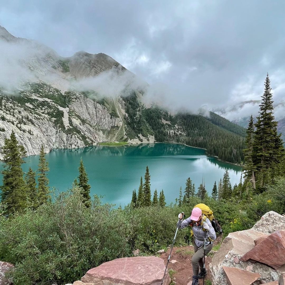 Woman wearing a flannel shirt, cap, and yellow hiking backpack with hiking poles walking the Four Pass Loop in Colorado on a foggy day | Photo by Instagram user @theearnestvoyager