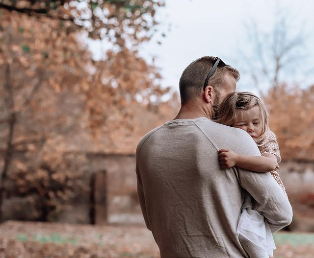 Father in beige sweater holding blonde daughter in his arms during fall photo shoot | Photo by Instagram user @bethanygracephotography