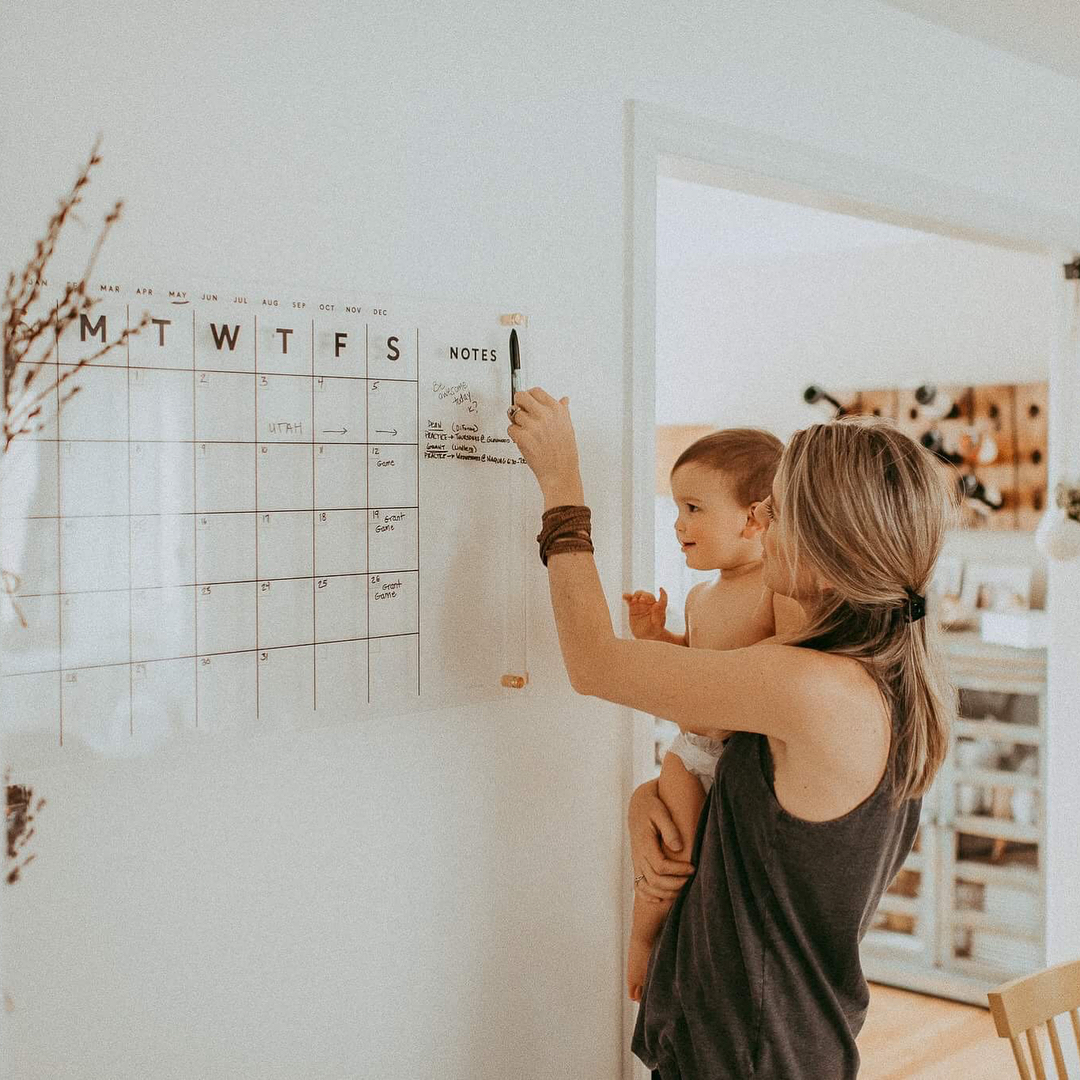 Mom holding baby while reaching for marker to write on clear acrylic calendar hung on the wall | Photo by Instagram user @elise_meader