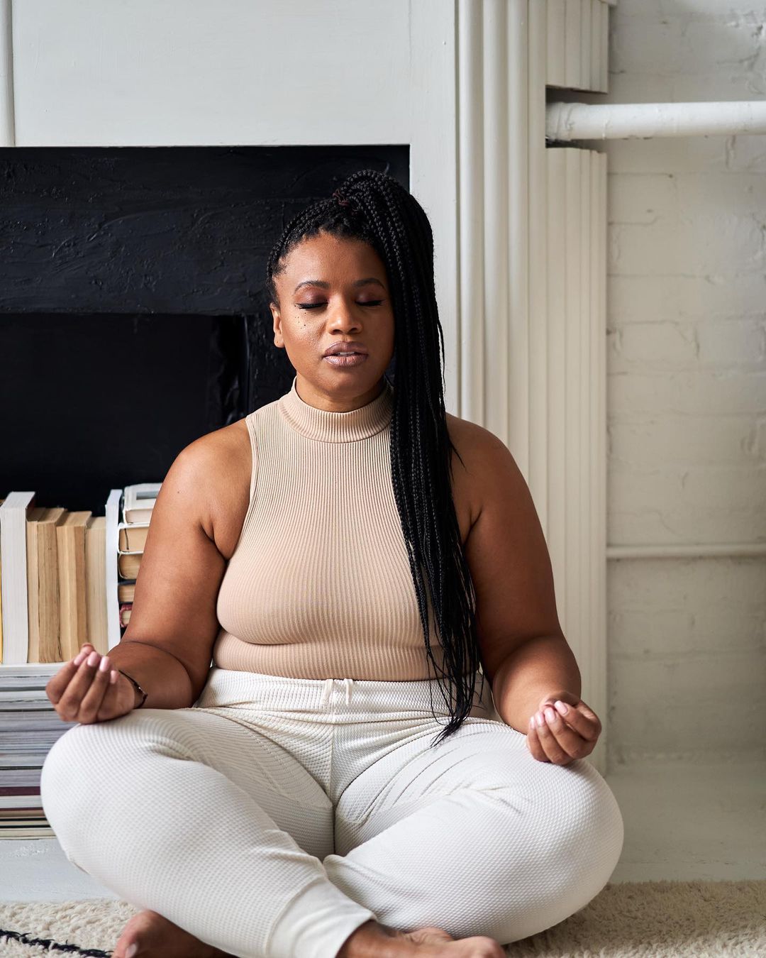 Woman in beige ribbed sleeveless turtleneck top and white ribbed sweatpants meditating in living room | Photo by Instragram user @rebeckahprice