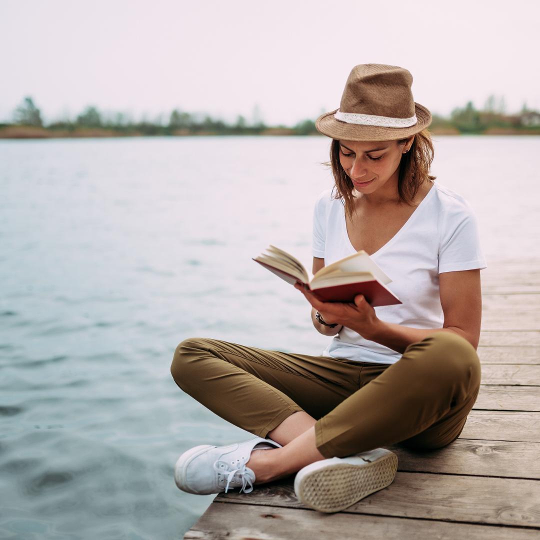 Woman wearing a tan fedora hat, white v-neck tshirt, brown jeans, and white tennis shoes sitting on a dock reading a book | Photo by Instagram user @joannamoranwellness
