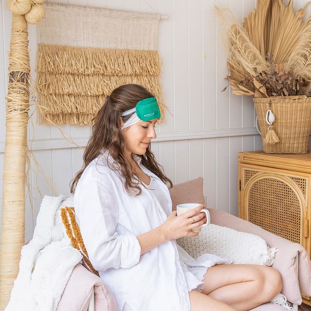 Woman closing her eyes wearing all white in a grass straw spa room holding a white mug and wearing white linen long-sleeve, shorts, and a green eye mask | Photo by Instagram user @noelleaustralia