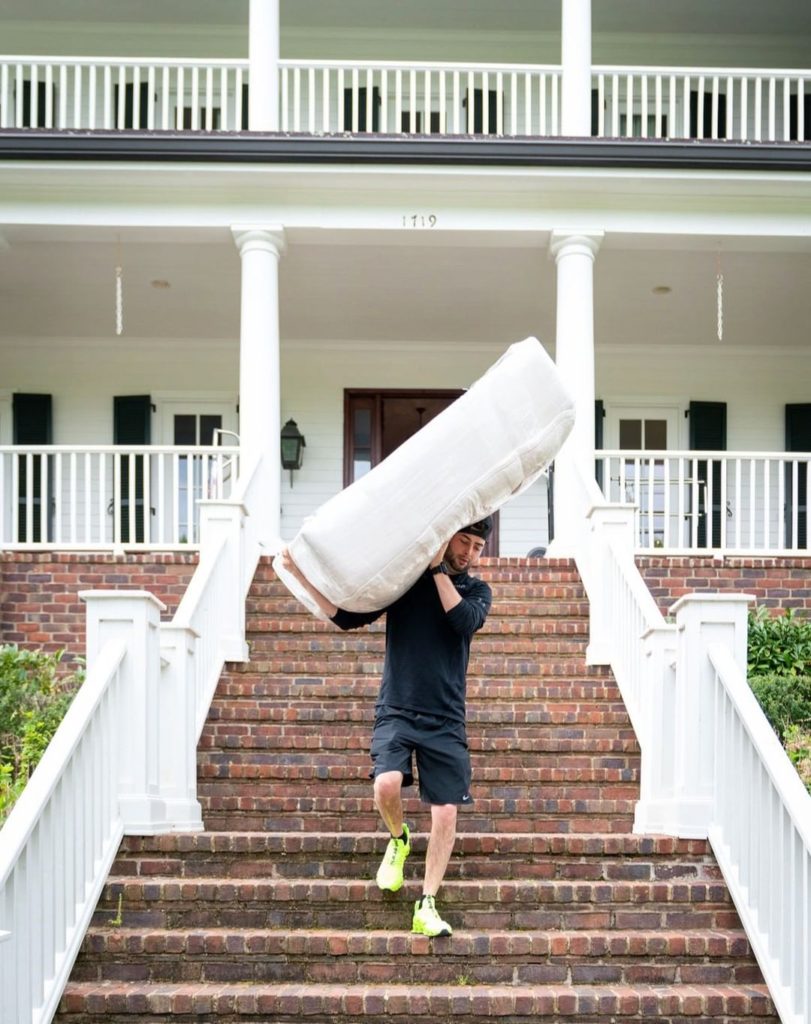 Man carrying a white mattress down a long series of brown steps in front of a federal style white home with white railings. Photo via Instagram user @blacktiemovingofficial