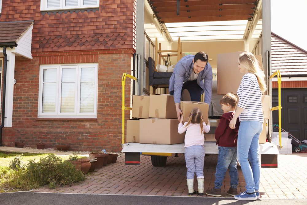 A man in a gray sweatshirt handing a box to a young girl while a son and mother watch, all at the back of a moving truck parked on the driveway of a brick house