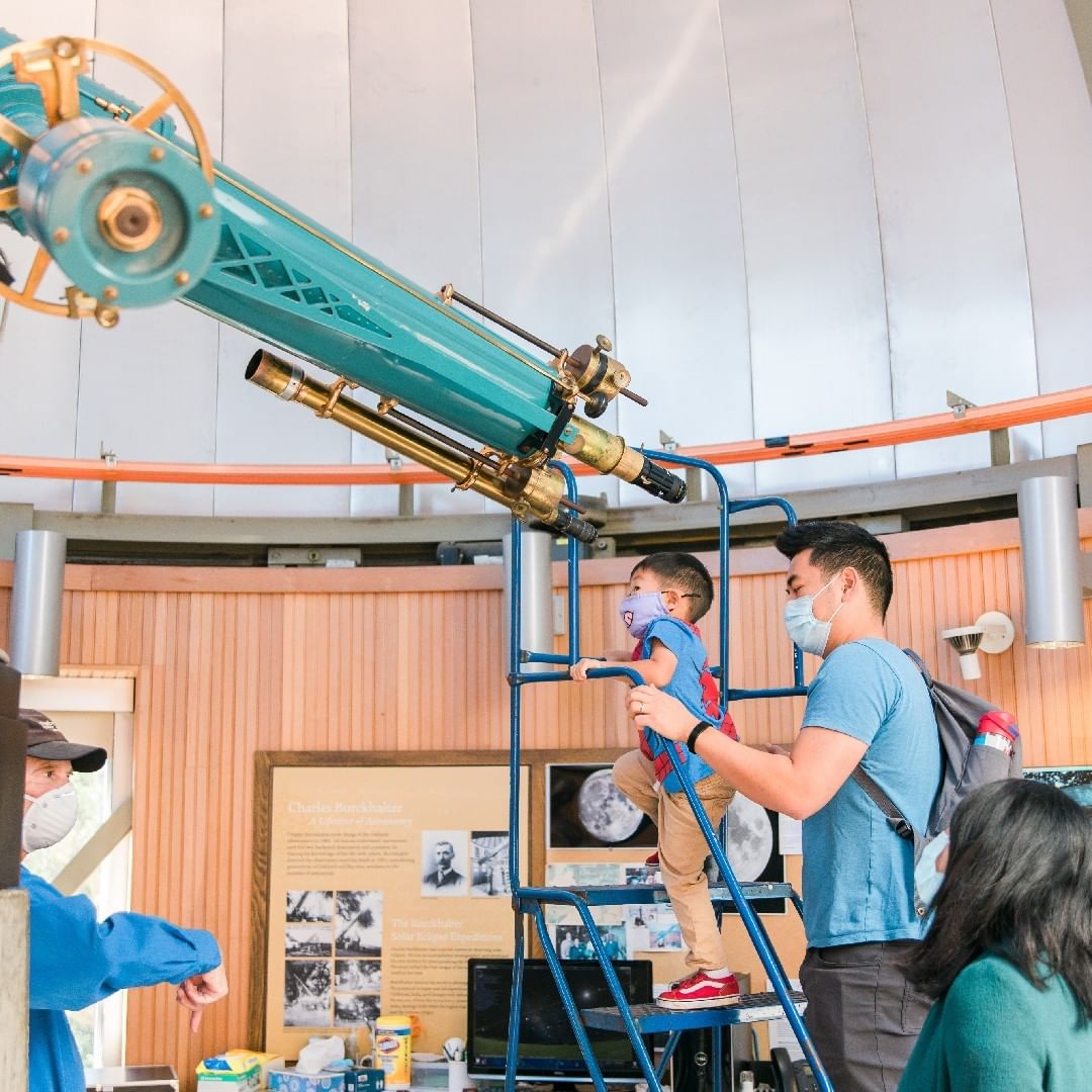 An adult man carrying a toddler so that he can look into a telescope in Chabot Space and Science Center. Photo by Instagram username @chabotspace