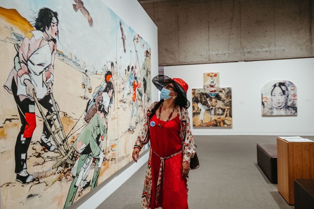 Lady in a red hat and dress walks by an art exhibition in Oakland Museum of California. Photo by Instagram username @oaklandmuseumca