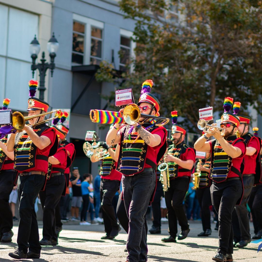Band members march while playing musical instruments like trombones and trumpets, wearing uniforms with rainbow colors during the Oakland Pride. Photo by Instagram username @oaklandpride22 