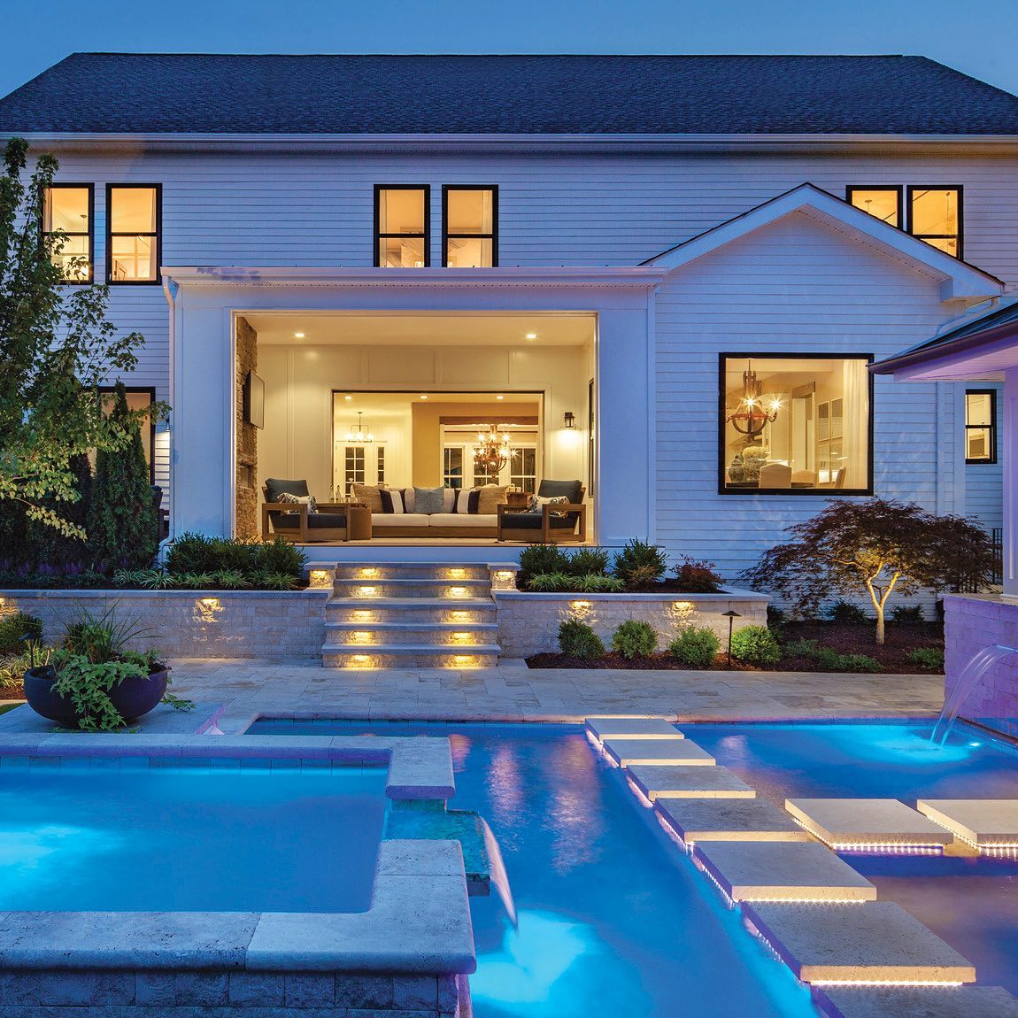 A look of a backyard with a lit pool in the foreground and a house in the background. @tollbrothers.