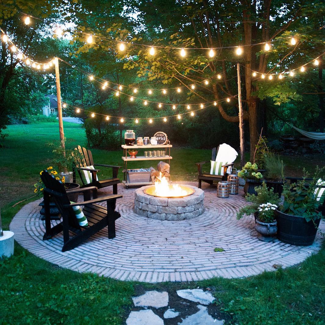 A backyard brick firepit area with lighting bar and chairs.