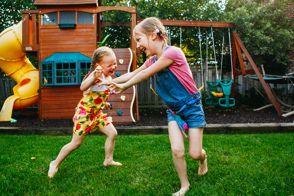 Two girls playing tag in front of wooden playset in grassy backyard