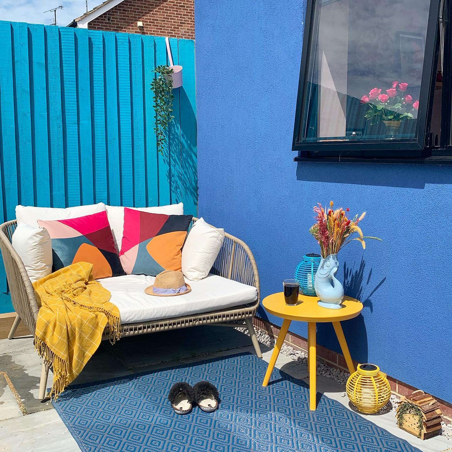 A backyard patio next to a bright blue fence decorated with a blue wall, and colorful pillows. Photo by Instagram user @carries_colourful_casa