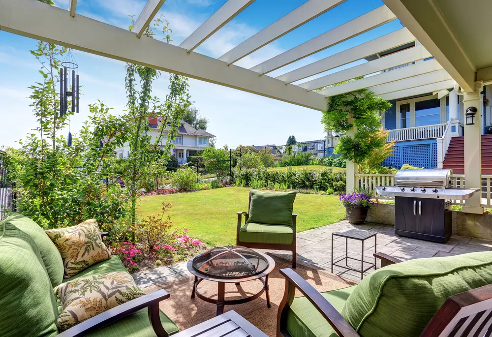 A sunny backyard patio with a small fire pit, grassy yard, and a pergola.