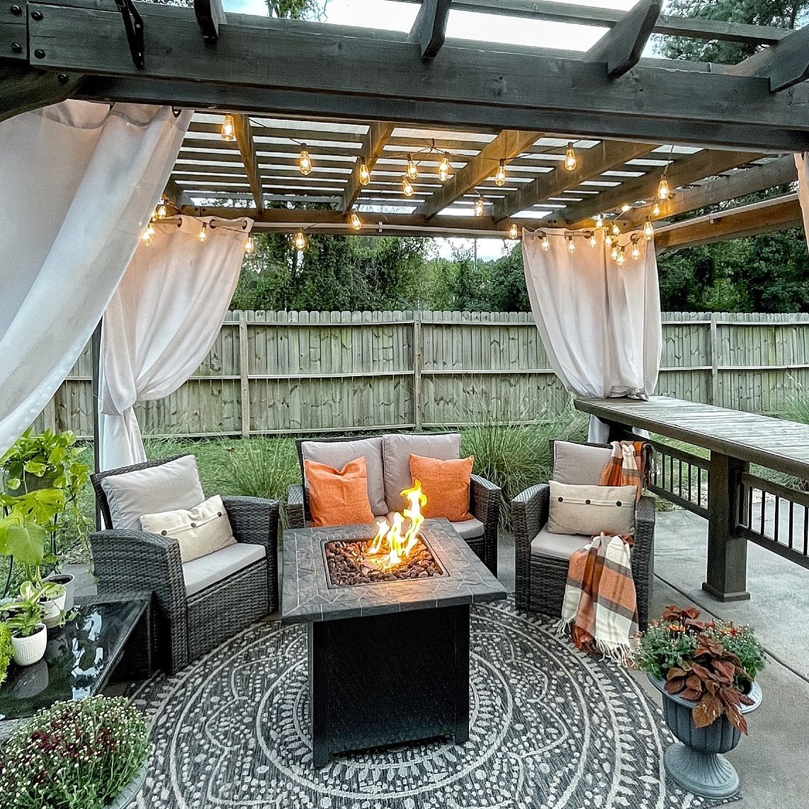 A backyard seating area with a fire pit in the center, beneath a pergola with curtains tied to its corners. Photo by Instagram user @shiplapshanty.