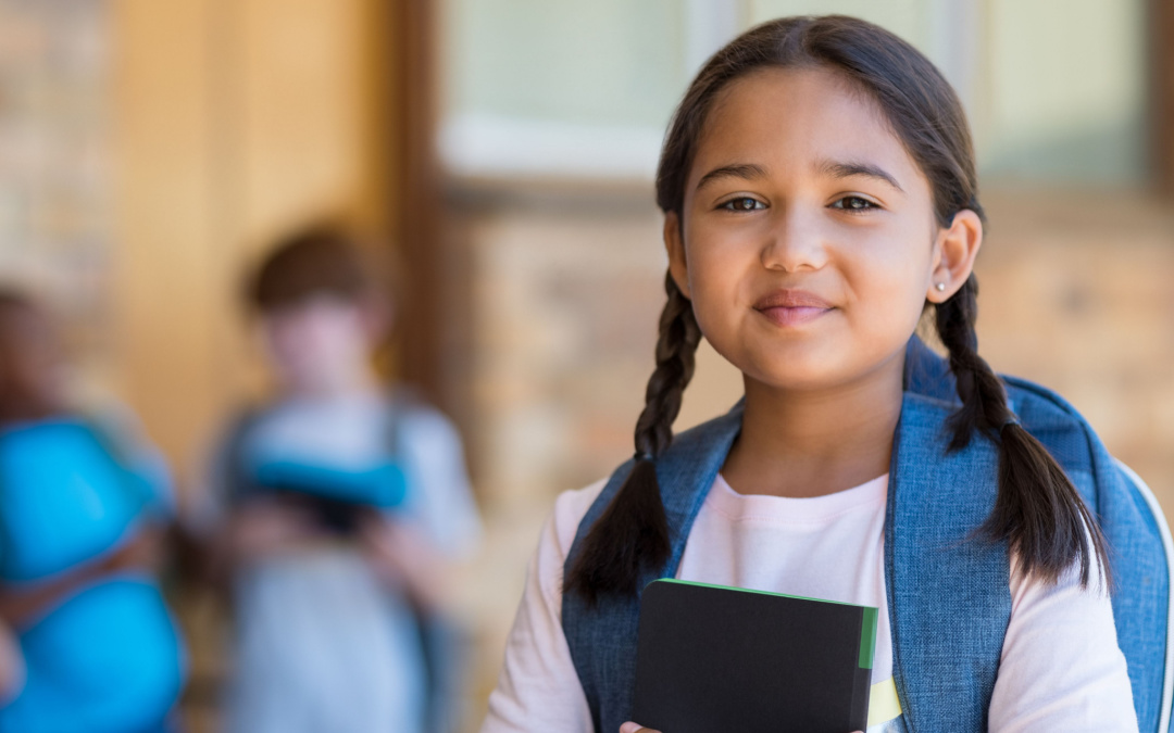 Young girl standing in school holding a notebook and carrying backpack.