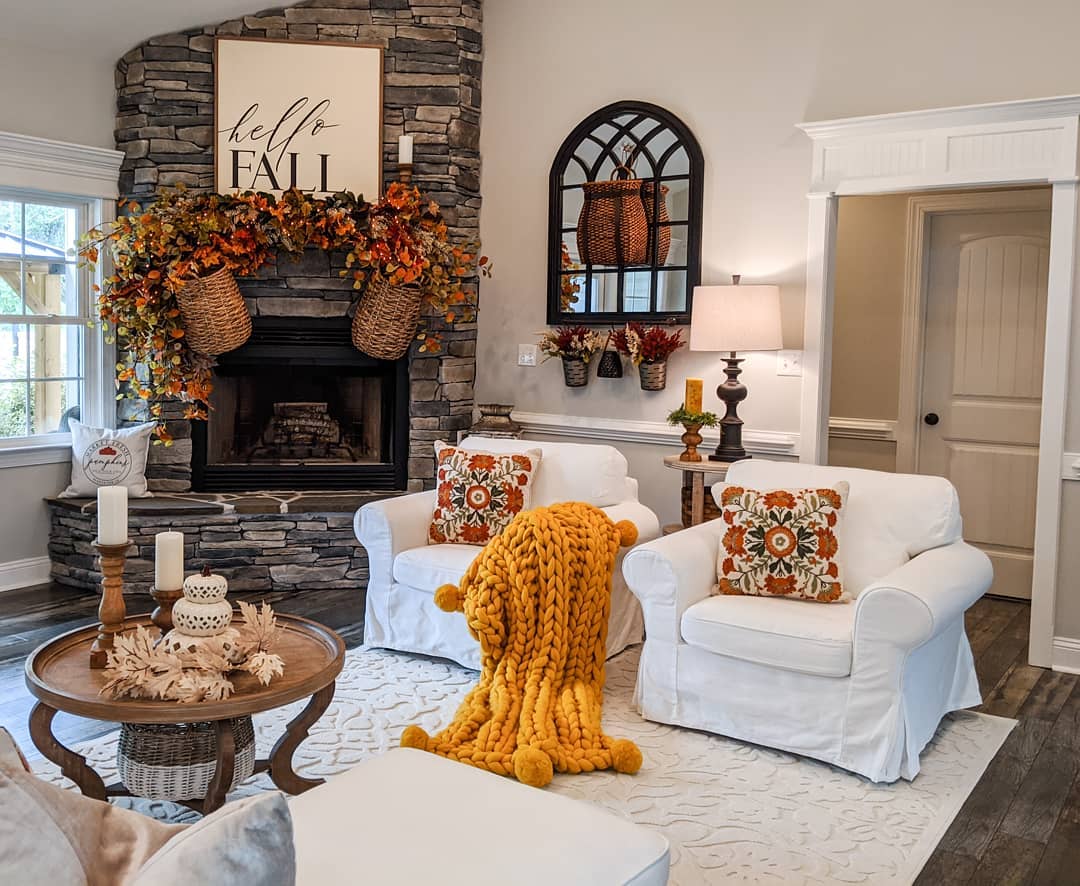 Living room area with two chairs, one of which is draped with a throw pillow, in front of a stone-facade fireplace decorated with fall foliage. Photo by Instagram user @bridgewaydesigns. 