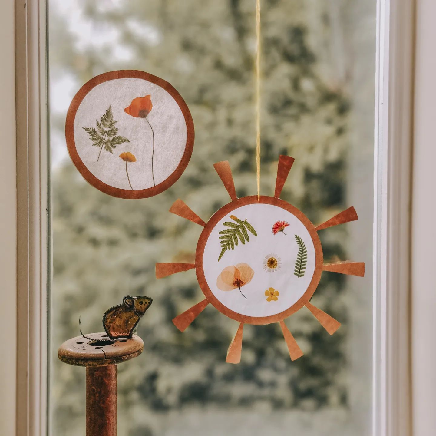 Circular crafts with the imprints of pressed flowers hanging against a window. Photo by Instagram user @kirstiecrafts.